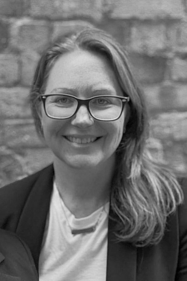 Black and white photo of a person wearing glasses and a blazer against a brick wall.